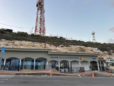 The image shows the exterior of a building with arched windows and doors, likely a visitor center or station, at the base of a rocky hill. A large red and white telecommunications tower stands to the left of the frame, and another smaller tower with a radome is visible on the hill to the right. The building has a sign with text in Hebrew and Arabic, including "WELCOME" in English. Several fences, safety nets, and traffic cones are present in front of the building, suggesting ongoing construction or maintenance. A pedestrian crossing is marked on the asphalt in the foreground. The sky is overcast. This scene is located in the city of Mate Asher Regional Council, Israel.