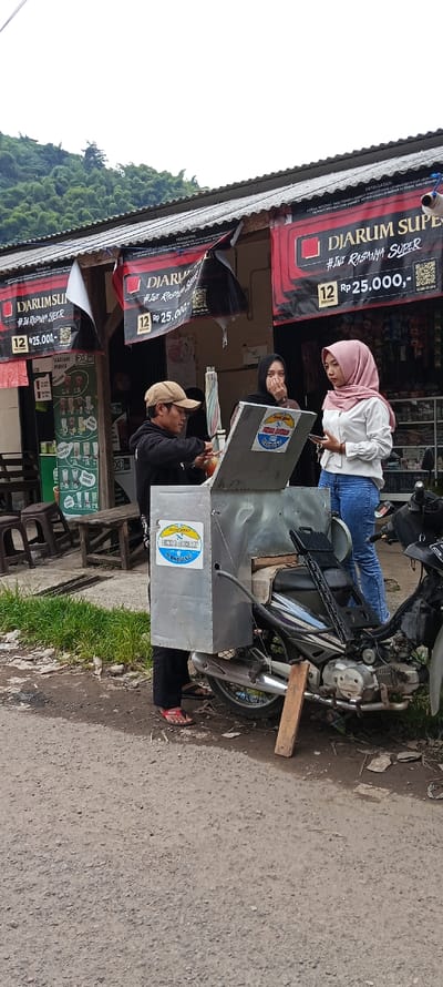 The image shows a street scene in Kutawaringin, Indonesia, located at coordinates -6.974 latitude and 107.519 longitude. Three individuals, two women and one man, stand in front of a store. One woman wears a white shirt and a light pink headscarf, while the other woman wears a gray shirt. The man wears a white shirt and a blue cap. The man holds a red cup and appears to be giving something to the woman in the gray shirt. Behind the man, there is a motorcycle with a gray metal box attached to the back. The store front displays numerous advertisements and signs, including a large sign for "Djarum Super." The store facade is made of concrete and the street is paved with gravel. The scene is set against a backdrop of green hills.