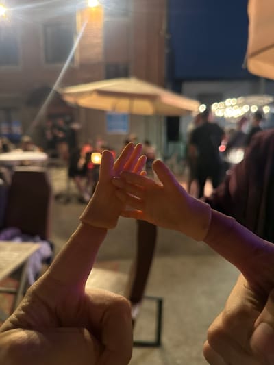 The image shows a close-up of two hands, one appearing to be a human hand and the other a prosthetic or doll's hand, positioned as if they are about to touch or interact. The human hand is in the foreground, with its thumb prominently visible. The prosthetic hand is further back, with its fingers outstretched. In the background, there is a blurred scene of an outdoor setting, possibly a cafe or restaurant area, with umbrellas, lights, and silhouetted figures of people. The lighting suggests it is dusk or evening. The overall scene conveys a sense of connection or gentle interaction between the two hands.
