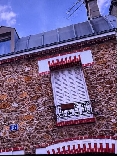 The image shows a close-up view of the exterior of a stone building with a blue sky in the background. The building has a pitched roof covered in dark gray metal, with a dormer window on the left side. There is a visible antenna attached to a chimney on the right side of the roof. The wall of the building is constructed from irregular stones of varying sizes and colors, primarily in shades of brown and gray, with mortar filling the gaps. A white-shuttered window is centrally located on the stone facade, framed by decorative red and white brickwork. A small black metal balcony with a planter box is attached to the window. A blue square sign with the number "15" in white is mounted on the stone wall to the left of the window. Red and white decorative brickwork is also visible along the top and bottom edges of the stone section of the building. The overall scene depicts an architectural detail of a building, possibly in a European city given the style of the architecture. There is no indication that this location is in Dubai, United Arab Emirates.
