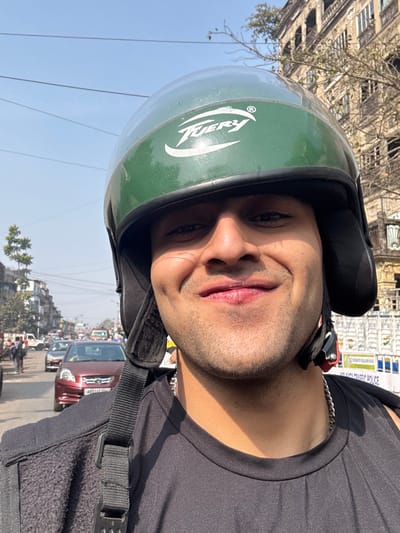 A man in a green helmet poses for a selfie on a city street. He wears a black t-shirt and a silver chain is visible around his neck. The background features a busy street with vehicles and buildings under a clear blue sky. The helmet has a white "Fiery" logo on the side. The man has a slight smile and is looking at the camera.