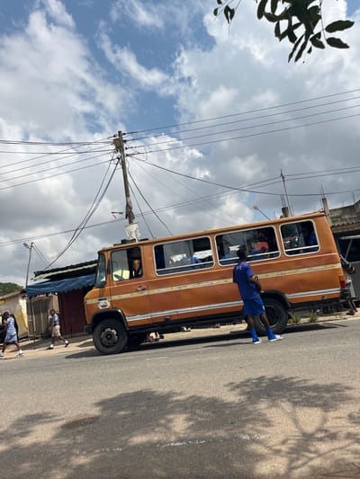 The image depicts a daytime street scene in a semi-urban area of Accra, Ghana, under a partly cloudy sky. The central focus is a weathered, orange-brown panel van or bus, likely a local 'tro-tro,' parked or stationary on the left side of a paved road. The vehicle features multiple rectangular windows along its side and appears to have occupants dressed in blue visible within.

Standing next to the passenger side of the orange bus, facing away from the camera, is a male individual dressed in a blue short-sleeved top, blue shorts, and blue and white athletic shoes. He holds a dark object in his right hand, extended downwards.

To the left of the bus, on a dirt and gravel shoulder adjacent to the road, two individuals are walking from left to right. The lead person wears a white and blue patterned dress and white shoes. The second person, slightly behind, wears a white and blue patterned shirt, light-colored shorts, and dark shoes.

In the background, on the left, are several single-story structures with blue fabric awnings or roofs, suggesting small shops or stalls. Further back, green foliage is visible. Dominating the upper left of the scene is a wooden utility pole heavily laden with numerous black electrical and communication wires extending in multiple directions. A street light fixture is mounted on this pole. On the right side of the road, behind the bus, are partially visible structures constructed from concrete blocks. The paved road surface shows faint markings and shadows from overhead trees, which are partially visible in the upper frame.
