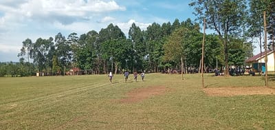 The image shows a grassy field in Bungoma, Kenya, at coordinates 0.553 latitude and 34.773 longitude. Five people are running in the field, away from the viewer. There are many trees surrounding the field and a small building is visible in the background on the left side. There are other people sitting in the shade of the trees on the right side of the image. The sky is partly cloudy. The grassy field is bare and there is a dirt path in the middle of the field. The people running in the field appear to be children, and they are wearing similar clothing. They are all running in the same direction, and their movements are fluid and effortless. The trees surrounding the field are tall and provide shade from the sun. The building in the background is a simple structure and appears to be a school building. The people sitting in the shade of the trees appear to be relaxing and enjoying the day. The overall atmosphere of the image is one of peace and tranquility.