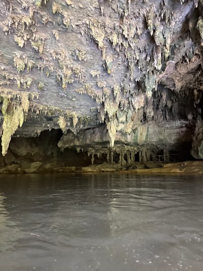 The image shows the interior of a cave with stalactites hanging from the ceiling and stalagmites rising from the ground. A body of dark, still water occupies the foreground. The cave walls are rough and textured, with some areas appearing damp and discolored. In the background, the cave opens up into darker recesses, and there are some wooden structures visible, suggesting a pathway or support. The overall atmosphere is dark and natural, characteristic of a cave environment. This scene is likely located within the caves of Ao Luek, Thailand, a region known for its karst landscapes and extensive cave systems.
