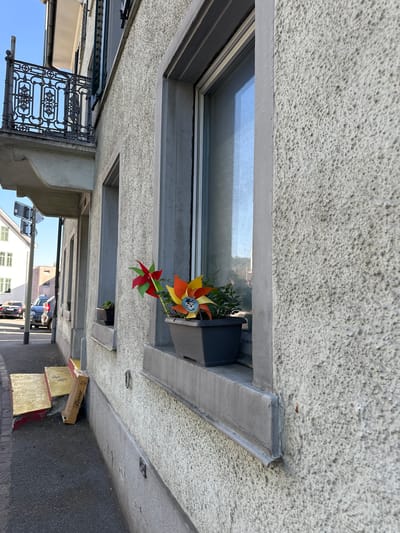 The image captures a section of a building's exterior in Wald (ZH), Switzerland, focusing on a window box containing two decorative pinwheels. The building has a textured, light-colored facade. One pinwheel is predominantly red and green, while the other features orange, yellow, and blue, with a ladybug design in the center. Both pinwheels are planted in a dark gray planter, which is situated on a window ledge. To the left of the window, a dark railing is visible, belonging to a balcony. Below the window, a sidewalk and a street are apparent, with cars parked and driving in the distance. A cardboard box leans against the wall near the sidewalk. The sky above is clear and blue.