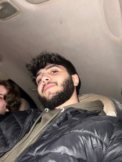 An image depicts two people inside a car. One is a young man with a dark beard, wearing a black puffer jacket over a hoodie. He's looking at the camera with a faint smile. The other person is partially visible at the bottom of the frame, with brown, wavy hair. The interior of the car is visible, including the roof and a beige car seat covered in plastic. The lighting is dim.