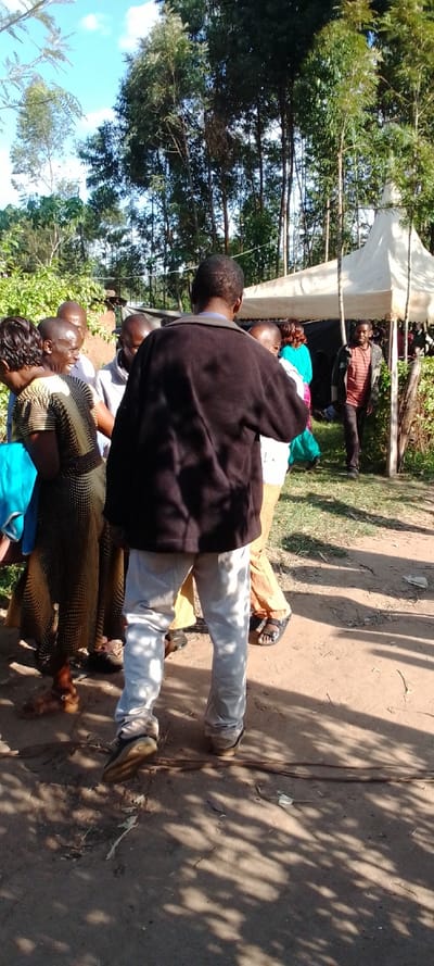 The image shows a group of people standing in front of a white tent in Kipkarren, Kenya, located at latitude 0.599 and longitude 34.957. The group consists of five adults and a young child. The people are wearing casual clothing. The person in the foreground, a man wearing a black jacket and white pants, appears to be the primary subject of the image. The people are standing on a dirt path, with trees and bushes in the background. The tent is partially obscured by vegetation. The scene appears to be an outdoor gathering or event. The sun is shining. The image is taken from a low angle and slightly off-center, with the primary subject positioned in the center of the image.  The image is well-lit, with a focus on the primary subject. The image is in focus and sharp, with a natural color palette.