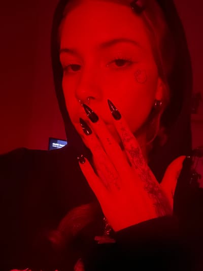 A close-up shot features a young person with blonde hair, wearing a black hooded garment. Their face is illuminated by red light, and they have a nose ring and a small Hello Kitty tattoo on their cheek. Their right hand, with long, sharp black nails, is raised to their face. The hand has intricate dark tattoos visible. The background is dark, with a faint glow from a screen in the lower left corner. There is no discernible location in Buenos Aires, Argentina.