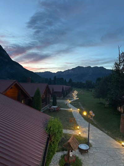 The image captures a serene twilight scene at a resort in Gusinje, Montenegro. In the foreground, the red corrugated metal roofs of several wooden cabins are visible, casting long shadows on the manicured lawn. A stone pathway winds through the property, illuminated by strategically placed outdoor lights and a decorative lamppost, creating a warm and inviting ambiance. The pathway leads past small, well-kept hedges and a rustic wooden wishing well with a decorative bucket. Further into the property, more cabins are visible, lined up along the pathway. In the midground, a heart-shaped sculpture stands on the grass, and a small cluster of outdoor seating with a table and chairs suggests a communal gathering spot. The background is dominated by the majestic, dark silhouettes of mountains, their peaks softened by the fading light. The sky above is a canvas of dusky blues and purples, with streaks of pink and orange from the setting sun, indicating the time of day is dusk. The overall atmosphere is one of tranquil beauty and peaceful seclusion, typical of a mountain resort.