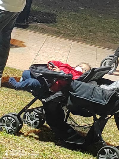 A baby in a red jacket and blue jeans lies in the left seat of a black double stroller. The baby has a pacifier in its mouth. Part of a person wearing dark pants and a light-colored shirt stands to the left. The stroller rests on a grassy area, with a paved sidewalk in the background. Another stroller wheel is visible to the right.