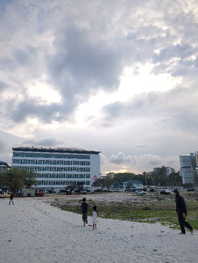 The image shows a white sandy beach with four people on it. The three children are standing on the sand, while an adult male wearing a dark jacket and black pants is walking away from the camera. The background is a cityscape with a large white building in the center, a few other buildings to the right, and a small, dense, green tree to the left. The sky is cloudy with a white sun shining through. This location is on the coast in Dar es Salaam, Tanzania, at coordinates -6.802 latitude and 39.289 longitude.