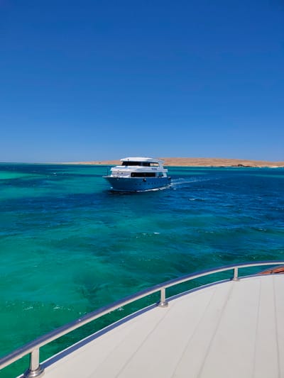 This is a landscape photograph taken from the perspective of a moving boat, likely in the Red Sea near Hurghada, Egypt. The image is dominated by a vast, clear blue sky above. Below the sky, the turquoise and deep blue waters of the sea are visible, with the water appearing exceptionally clear and shallow in some areas, revealing hints of coral reefs or sandy bottoms. A white and blue yacht is the central subject, sailing away from the viewer, leaving a visible wake. In the distance, a low-lying, arid coastline stretches across the horizon, characterized by sandy, barren hills. The foreground of the image shows the white deck of the boat from which the photograph is taken, complete with a polished chrome railing. The scene evokes a sense of travel, leisure, and the natural beauty of a coastal environment.