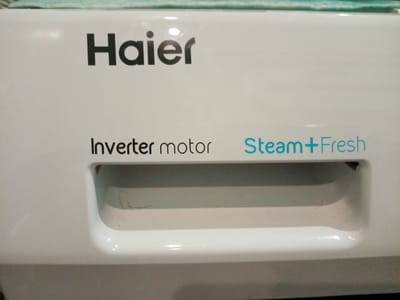 The image shows a close-up of a white Haier washing machine. The brand name "Haier" is printed in black at the top. Below that, the phrase "Inverter motor" is printed in black, and to the right, "Steam+Fresh" is printed in blue. A handle is located below this text. The image provides no information to infer a location in Elektrostal, Russia.