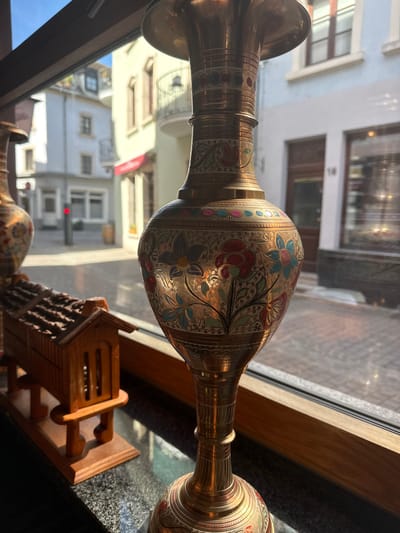 A still life photograph displays two ornate, brass vases in front of a window overlooking a street scene, likely in Grevenmacher, Luxembourg. The vases feature intricate, colorful floral designs. A small wooden object, possibly a decorative piece, is also present on the windowsill. The window reflects buildings across the street, providing a view of the urban landscape. The overall lighting is bright, with sunlight streaming through the window.