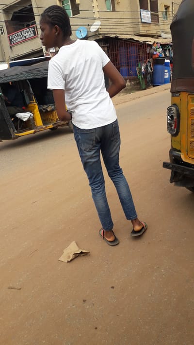 A woman is standing on a street in Nkpor, Nigeria. She is wearing a white t-shirt, blue jeans, and sandals. A yellow vehicle is partially visible in the background. A piece of cardboard lies on the street. Buildings and power lines are visible in the background, along with what appears to be a shop and its contents.