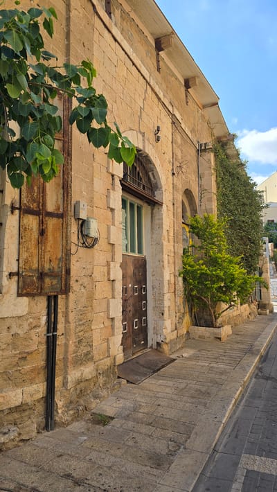 A side view of an old, weathered stone building with a metal door and a tree in a planter is captured on a sunny day in Tel Aviv, Israel. The building's facade is constructed from large, roughly cut stone blocks, with a noticeable patina of age and some areas of discoloration. Above the stone, the upper part of the building features a lighter-colored, possibly painted, section with exposed wooden beams supporting a sloping roof.
The entrance is marked by a dark, metallic door with a grid-like pattern and several square panels. A metal ramp leads up to the door, and a small, diamond-patterned metal mat is placed in front of it. To the left of the door, a rusty metal panel is affixed to the wall, with electrical conduits running vertically alongside it. A grey electrical box is mounted on the stone wall above the rusty panel.
A green tree with broad leaves is situated to the right of the entrance, planted in a rectangular stone planter. The building is bordered by a paved walkway constructed from large, irregular stone slabs, which slopes downwards towards a street paved with smaller, darker stones. The sky is a vibrant blue with scattered white clouds. The overall scene suggests a historical urban setting in Tel Aviv, characterized by its distinctive architecture and Mediterranean climate.