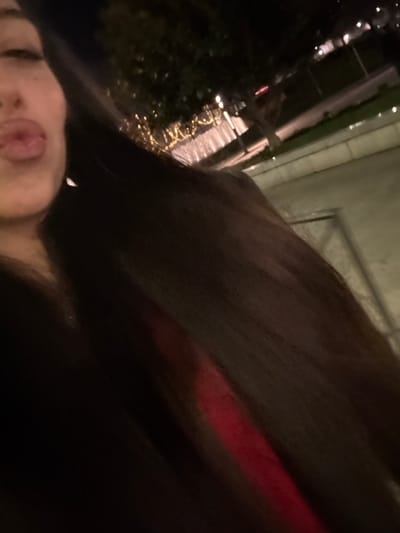 A selfie-style image, dimly lit, features a person's face in the lower left corner, with their tongue sticking out. The background appears to be nighttime outdoors, with decorative lights and possibly a building facade visible. Most of the frame is filled with a dark, blurry mass that could be hair or a garment. A hint of red suggests fabric or lighting. The overall composition is tilted and unbalanced.