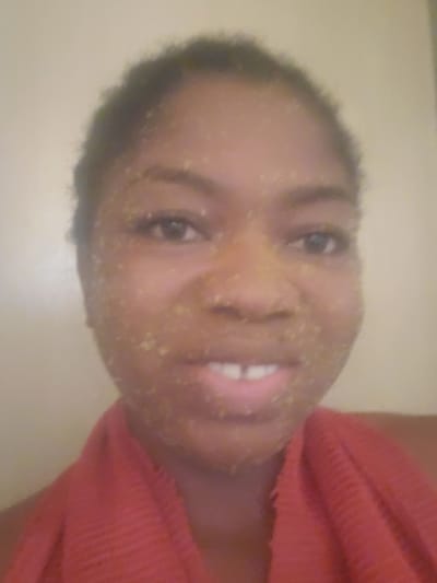 A frontal, close-up portrait depicts an adult individual with dark skin and short, dark, curly hair, smiling directly at the camera. Their face is entirely covered with numerous small, light yellowish-green, granular particles, consistent with a facial scrub or mask. The person's mouth is open in a smile, revealing a visible gap between their two upper front teeth. A red, textured fabric, likely a towel or garment, is draped around their neck and shoulders. The background is a plain, uniformly light beige or off-white wall, indicating an indoor setting. The image provides no visual cues to determine a specific geographic location, such as the city of Kakau, Nigeria.