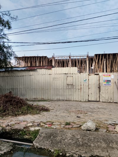 The image depicts a construction site in Moba, Nigeria. A corrugated metal fence, showing signs of rust and wear, encloses the site. Above the fence, the wooden scaffolding of a multi-story building under construction is visible, supported by numerous vertical wooden poles. The sky above is partly cloudy with several power lines strung across it. In the foreground, a pile of debris, consisting of branches and roots, lies on a cobblestone ground. Adjacent to the debris is a narrow channel of murky water, with concrete slabs at its edge. On the metal fence, there are several signs, including one indicating "RESTRICTED AREA CONSTRUCTION SITE" with emergency contact information. There are no people visible in the image.