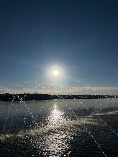The image depicts a body of water, likely Ternopil Pond, under a clear blue sky with the sun prominently visible. A fountain is in operation, with arcs of water droplets creating a sparkling effect against the sunlight. In the distance, a silhouetted tree line borders the water, with what appears to be a city skyline and a radio tower visible atop the hills. The water itself is dark and reflects the sunlight, with smaller splashes and ripples evident. The overall impression is of a bright, sunny day with a recreational or scenic setting.