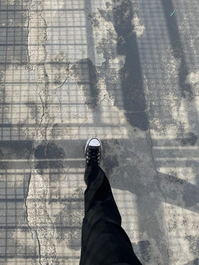 The image depicts a person standing on a cracked, weathered concrete surface in an outdoor location. The person is wearing black pants and a black and white Converse sneaker. The surface is marked by shadows of a wire mesh or grid-like structure, likely a fence or railing above. The background surface appears to be a road or sidewalk. A green dental floss pick is visible in the upper left of the image. No further details are available to indicate that the photo was taken in Nogent-sur-Oise, France.