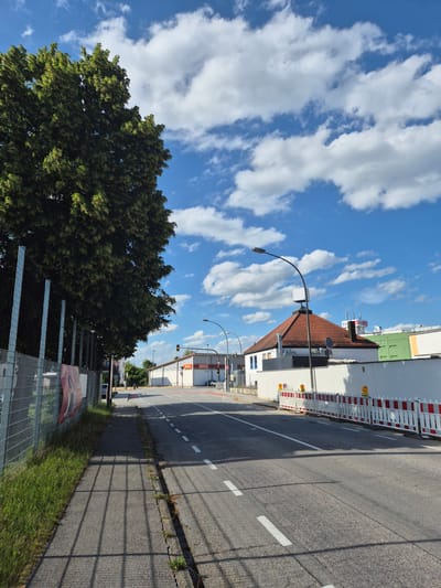 The image depicts a street scene in Landshut, Germany, on a sunny day with scattered clouds. On the left side of the road, a chain-link fence with metal posts runs alongside a grassy area and a sidewalk. A large tree with dense green foliage partially obscures the sky. The road itself is paved and marked with white dashed lines, indicating traffic lanes.
On the right side of the road, several buildings are visible. One building has a prominent red-tiled roof and a white facade, with a street lamp in front of it. Another building, likely a commercial establishment, features the "NORMA" logo on its facade and has a traffic light in front. Further down the street, construction barriers with red and white stripes are set up along the side of the road, suggesting ongoing work. The overall environment appears to be an urban or suburban setting with a mix of residential and commercial properties. There are no people visible in the image.