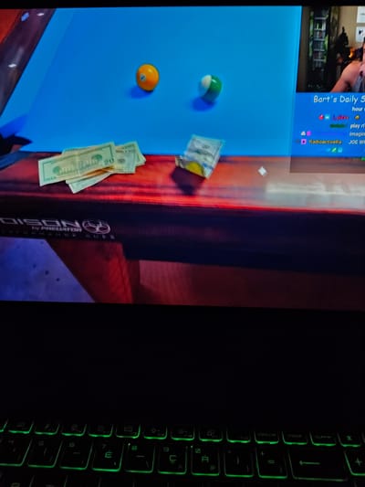 This image shows a close-up view of a computer screen displaying a digital simulation of a pool table with two billiard balls and a stack of US dollar bills on a red surface. The background of the screen is a vibrant blue, representing the pool table's felt. To the right of the pool table simulation, a portion of a Twitch stream is visible, featuring a person in the upper right corner and a chat interface with user names and messages. Below the pool table, a portion of a keyboard with illuminated green keys is visible. The scene is forensic and matter-of-fact, depicting digital objects on a screen and the physical interface of a keyboard. There is no indication of the city of Versailles, France, in this image.