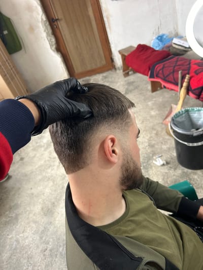 This image depicts a haircut service taking place in an indoor, semi-finished space located in the city of Hvostiane, Bulgaria. A male individual, seated and viewed from the rear-right, is receiving a haircut. He has short, dark hair with a distinct skin fade on the sides and back, tapering from bald to longer on top, and a trimmed beard. A hand, gloved in black nitrile, is positioned on the top of his head, with the sleeve of a red and navy blue striped garment visible on the arm. The environment features light-colored, textured walls with visible wear and cracks, and an unpolished light grey concrete floor. In the background, from left to right, are a partially visible cardboard box, a closed brown wooden door with a dark handle, a small wooden stool topped with a red and black patterned blanket, a blue plastic bag, a wooden-handled brush or broomstick, a black plastic bucket with a light liner, a small white object on the floor, and a circular white light source (ring light) partially visible on the right.