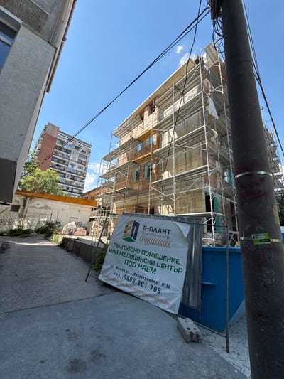 In Shumen, Bulgaria, a construction site is visible. The scene features a multi-story building undergoing construction, surrounded by scaffolding. A large banner with Bulgarian text, advertising commercial or medical spaces and providing contact information, is prominently displayed. The banner includes the address "gr. Shumen ul. „Industrialna“ №30" and a phone number, "0888 201 706," along with a website and email address. A tall utility pole with numerous wires stands to the right of the frame, and other buildings, including a residential high-rise and a lower structure with graffiti, are visible in the background. The environment suggests an urban setting under a clear blue sky.