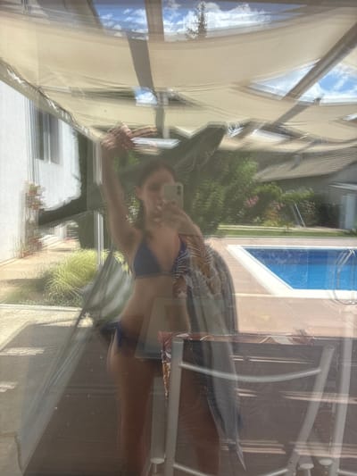 A young woman in a blue bikini and a patterned cover-up is reflected in a window, holding a phone to take a selfie. She is standing on a wooden deck or patio, with a swimming pool and lush greenery in the background. The scene appears to be a private residence, possibly in the city of Bulgarovo, Bulgaria, given the architectural style and vegetation. The reflection suggests it is daytime, with sunlight filtering through a canvas awning overhead.