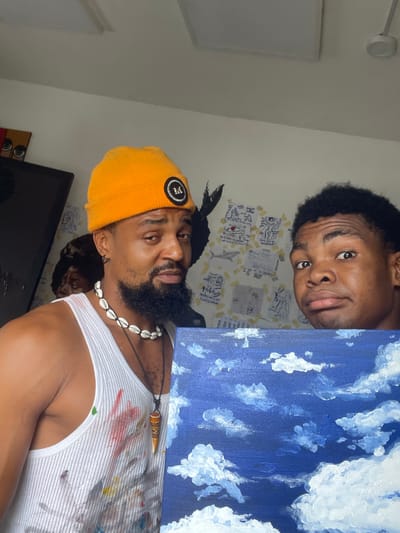 The image depicts two men and a painting. The man in the foreground is wearing a yellow beanie, a white tank top with paint splatters, and a shell necklace. He has a beard. He is holding a painting of a blue sky with white clouds. The man in the background has short black hair and is looking at the camera. The background wall is decorated with drawings or notes taped to the wall. The location is likely indoors, possibly in Ado, Nigeria. The light is dim, and the orientation of the image appears rotated.