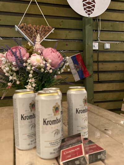 In Kochetinskiy, Russia, on a wooden table, are four cans of Krombacher beer and two packs of Chapman cigarettes. A bouquet of pink and white flowers sits behind the cans. A Russian flag is draped on a wooden fence in the background. There is also a decorative hanging object filled with wine corks and a circular decoration featuring a pine cone motif.
