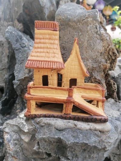 A small, intricately designed ceramic house, resembling a miniature pagoda, rests on a textured gray rock formation. The house is predominantly light brown with darker brown accents, featuring layered roofing that mimics thatched tiles and small, dark openings suggesting windows and a doorway. A raised platform with railings and a ramp connects different sections of the house. The background is softly blurred, showing more rock formations and a hint of colorful lanterns and greenery, suggesting an outdoor setting. This scene is likely located in Da Lat, Vietnam, a city known for its unique architecture, French colonial influences, and abundant natural beauty, often incorporating rock gardens and artisanal crafts into its public spaces and private residences.