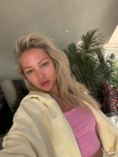 A blonde-haired woman with fair skin poses for a selfie. She is wearing a pink top and a cream-colored jacket. In the background, there are green plants and part of a building. Based on the style of architecture and vegetation, the photo was likely taken in El Gouna, Egypt.