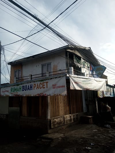 A two-story building with a shopfront banner that reads "Rumah Buah Pacet" (Pacet Fruit House) is visible. The building appears worn, with visible deterioration on the façade. Power lines run densely above the structure. The shopfront is partially constructed of wood and has a concrete base. The street level and surrounding areas are dimly lit and appear somewhat dirty, suggesting a less affluent neighborhood.