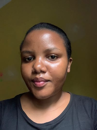 A close-up portrait shot of a Black woman with short, dark hair. She is wearing a black top and is looking at the camera with a slight smile. The background appears to be a wall with an olive-green hue. There is no information to suggest that this photo was taken in Dar es-Salaam, Tanzania.