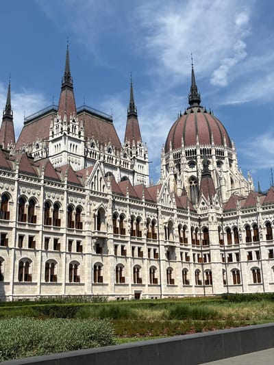 The image shows a detail of the Hungarian Parliament Building in Budapest, Hungary. The building is a grand Gothic Revival structure with ornate facades, multiple spires, and a prominent dome. The exterior is made of light-colored stone, and the roofs are tiled in red. Several arched windows with dark shutters are visible along the facade. In the foreground, there is a neatly landscaped area with green bushes and tall grasses. The sky above is blue with scattered white clouds.