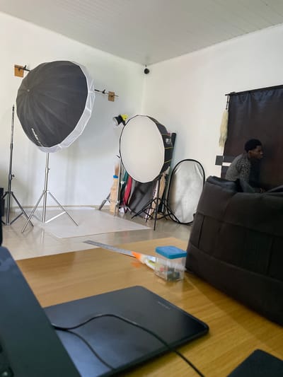 In a room that appears to be a photo studio, possibly located in Durumi, Nigeria, a person is seated. The individual, a young Black male with short dark hair, is wearing a grey, long-sleeved, patterned shirt. He is positioned facing right, looking away from the camera, and is seated near a dark backdrop.
The studio is equipped with various photography lighting equipment. To the left, a large softbox umbrella, branded "Godox," is mounted on a stand. Another softbox, smaller and circular, is visible to the right of the first one, also on a stand. A silver-sided reflector is placed between the two softboxes. Various light stands and photographic accessories, including what looks like broomsticks, are also present.
In the foreground, a wooden desk is visible, covered with some items. A laptop or tablet is partially in view on the left side of the desk. A ruler and a small clear plastic container with a blue lid, containing small colorful objects, are also on the desk. To the right, a large black bag or case rests on the floor. The walls are white, and the ceiling appears to be paneled. The floor is tiled with a light-colored grid pattern. A white sheet of paper or thin mat is spread on the floor in front of the lighting equipment. A security camera is mounted high on the wall.