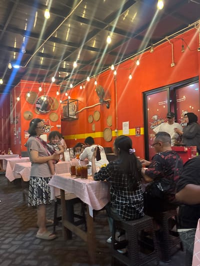 The image captures an evening scene inside a casual restaurant, identified as "Garasi Sidoarjo - Restoran Ayam Bakar," located in Sidoarjo, Indonesia. The establishment features vibrant orange-red walls, decorated with multiple circular woven bamboo winnowing trays of varying sizes and a large mirror. A dark ceiling is illuminated by numerous exposed Edison-style light bulbs strung in rows, casting visible light streaks. The floor is composed of light-colored brick pavers.
In the left foreground, a female subject wearing glasses, a light brown short-sleeved top, and a patterned knee-length skirt stands holding a small child (toddler) in a patterned baby carrier. Both wear light-colored footwear. In the mid-ground, a group of individuals is seated at a square wooden table draped with a pink and white checkered tablecloth. Three glasses containing a dark liquid and straws are visible on this table. One person, wearing a black and white plaid shirt, is seated with their back to the camera. Another male, wearing a black and red patterned shirt and glasses, is seated facing left, with a black Reebok bag visible on his lap. Further back at an adjacent table, a person in a white long-sleeved shirt is partially visible. In the background, behind a glass partition featuring a red chicken logo and "Garasi" text, two individuals are standing: a male wearing a white t-shirt, black baseball cap, and sunglasses, and a female wearing a dark hijab. A wall-mounted fan is situated on the right wall. Exposed electrical conduits and wiring are visible throughout the space.