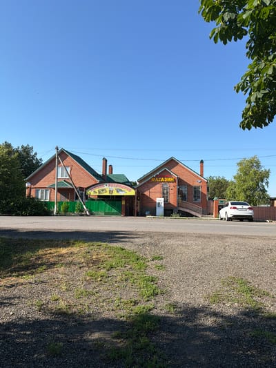 The image shows a sunny day scene in front of two adjacent brick buildings on a street in Leningradskaya, Russia. The building on the left has a green metal gate and a sign that reads "ЖИВОЕ • РАЗЛИВНОЕ" with images of fish. The building on the right has a sign that says "ПРОДУКТЫ МАГАЗИН". A white car is parked in front of the building on the right, and a white vertical machine is next to the entrance of the building on the right. Trees and a fence are visible in the background. The sky is clear blue with a few distant birds. The ground in the foreground is covered with gravel and sparse grass.