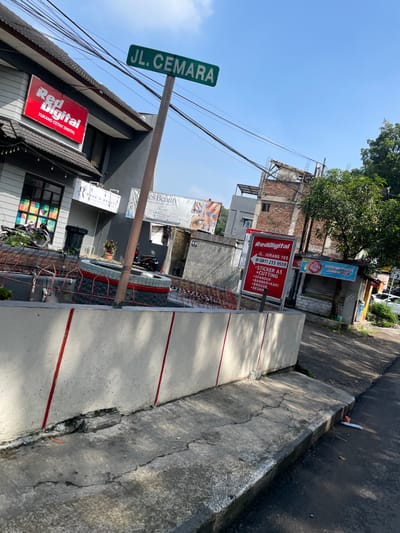The image captures a street scene in Bandung, Indonesia, at the coordinates -6.885 latitude and 107.601 longitude. The street, named "JL. CEMARA", is lined with various businesses and buildings. A concrete curb separates the street from the sidewalk. A grey concrete wall with red vertical stripes runs along the sidewalk, indicating a possible boundary or property line. A business called "RedDigital" is prominent on the left side of the image, showcasing its sign with the text "TUKANG CETAK DIGITAL". There is a small sign for another business named "Es Beauty" displaying nail art designs. To the right of the image, a second "RedDigital" sign is visible, along with an unidentified business sign advertising "STIKER A3 CUTTING". Motorcycles are parked in front of "RedDigital" on the left. The scene is captured from a perspective looking down the street towards a small, partially obscured building. The sky is blue with no clouds, and the surrounding environment is typical of an urban area in Indonesia.