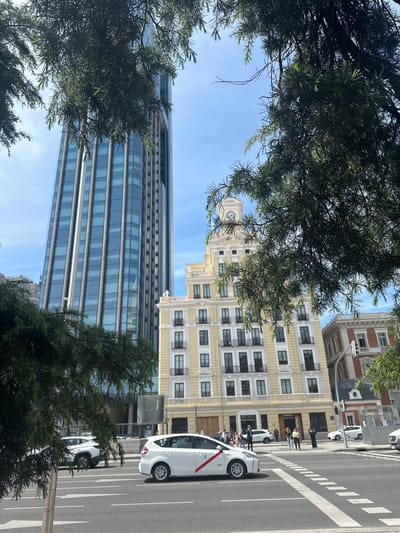 The image captures an upward-looking street scene in Madrid, Spain, framed by dense green foliage of trees in the foreground and along the sides. The clear blue sky is visible between the branches.

In the middle ground, a modern, tall skyscraper with reflective blue-tinted windows stands prominently. To its right, an older, ornate building with a distinctive clock tower at its top is visible, showcasing traditional European architecture. Other historical-style buildings with multiple windows and balconies are seen further in the background.

On the right side of the frame, a street with a pedestrian crossing marked by white zebra stripes is visible. Several white taxis, characteristic of Madrid with a red stripe along the side, are on the road. One taxi, closest to the foreground, has the number "9079" visible on its rear. A few people are standing and walking on the sidewalk adjacent to the buildings, engaged in typical urban activity.

The scene is illuminated by bright daylight, casting distinct shadows on the zebra crossing and highlighting architectural details. The blend of modern and traditional architecture, along with the specific taxi design, provides clear location cues for Madrid.