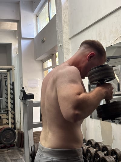 The image shows a person with short, light brown hair performing a weightlifting exercise with a dumbbell. The person is shirtless, wearing grey athletic shorts. The dumbbell is heavy, with multiple weight plates stacked on it. The person is holding the dumbbell with their right hand, positioned behind their head. The background shows a gym setting with weight racks and other gym equipment. The lighting suggests it might be daytime, with natural light coming from windows visible in the upper left of the image. The overall scene appears to be an indoor gym, possibly in a slightly rustic or older facility, given the visible concrete and painted ceiling with some wear and tear. There are no discernible emotions visible on the person's face due to the angle and focus of the shot. Text is visible on a white paper attached to the wall behind the person, but it is too blurry to read. The location context provided is Shumen, Bulgaria.
