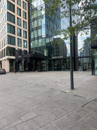 The image depicts the modern architecture of the NYX Hotel and Varso Place in Warsaw, Poland. In the foreground, a vast, grey tiled plaza stretches out. A solitary dark pole stands in the middle of the plaza, casting a subtle shadow. To the left, a black car with a license plate visible is parked. The NYX Hotel entrance is adorned with several large black flags bearing the hotel's logo. Further to the right, the entrance to Varso Place is visible, marked by prominent signage. The buildings are characterized by extensive glass facades, reflecting the sky and surrounding structures. A few individuals are present in the scene, with one person standing near the hotel entrance and another near the Varso Place entrance. A tree with green leaves branches out from the upper right corner of the frame, partially obscuring the building's facade.