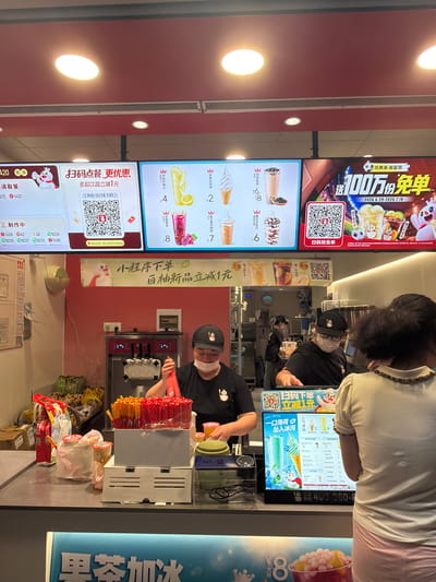 This image depicts a brightly lit fast-food establishment, likely a dessert or beverage shop, with a prominent display of menu items and promotional material. Two employees, wearing black shirts and caps, are visible behind the counter. One is actively preparing a beverage, holding a red dispenser. The other, partially obscured, is also behind the counter. In the foreground, a person in a white top and pink skirt is seen from the back, looking at the menu.
The setting is indoors, with overhead lighting contributing to the overall brightness. The walls are a vibrant red. Digital menu boards display images of drinks like milk tea and ice cream, along with prices. One large banner at the top advertises a "1 million free orders" promotion, valid from June 29, 2026, to July 18, 2026, encouraging customers to scan a QR code. Another digital screen offers a discount for ordering via a mini-program.
Various objects are visible on the counter, including dispensers for soft-serve ice cream, cups, and packaged snacks. The overall atmosphere suggests a busy service environment. Visible text includes promotional slogans, menu item names and prices, dates, and instructions for ordering, primarily in Chinese. The presence of QR codes indicates a reliance on digital payment and ordering systems. The location context is identified as Jiangning District, China.