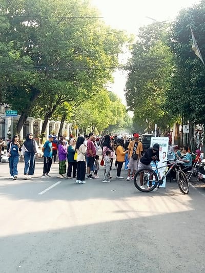 The scene is on a paved road in Pati, Indonesia (latitude: -6.753, longitude: 111.04), under a partly cloudy sky, with a group of about 25 people standing on the road, facing a large building on the right side of the road. There are several trees on both sides of the road. Two people are walking away from the group towards the left side of the road. A bicycle sits in the foreground, on the right side of the road. Some of the people are wearing traditional clothing. There are cars parked on the side of the road.  The large building on the right side of the road is a business with signage. One of the people is using a cell phone.  The people are interacting with each other and appear to be enjoying themselves. The building in the background appears to be a business with signs that have several different colors and different lettering. The street is a two-lane street, and the road is paved.  There are no parked cars on the left side of the road. There are several power lines running along the top of the image.  The image has a bright and cheerful tone.  The group appears to be in a public place.