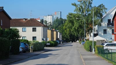 The image depicts a residential street in Linköping, Sweden, on a clear, sunny day. The street is lined with houses and apartment buildings, interspersed with lush green trees and hedges. In the foreground, a paved road curves gently to the right, with a sidewalk on the left. Several cars are parked along the street and in driveways.
On the left side of the street, a two-story house with a red brick facade is partially visible, followed by a light-colored apartment building with a red-tiled roof. Beyond this, a row of yellow and cream-colored apartment buildings continues down the street. In the distance, several taller apartment buildings are visible against a blue sky.
On the right side of the street, a white apartment building with balconies is prominent, and further down, a red-colored building is partially visible. A white car is parked on the right side of the road. The scene is peaceful and suburban, with no people visible. The environment suggests a well-maintained residential area.
