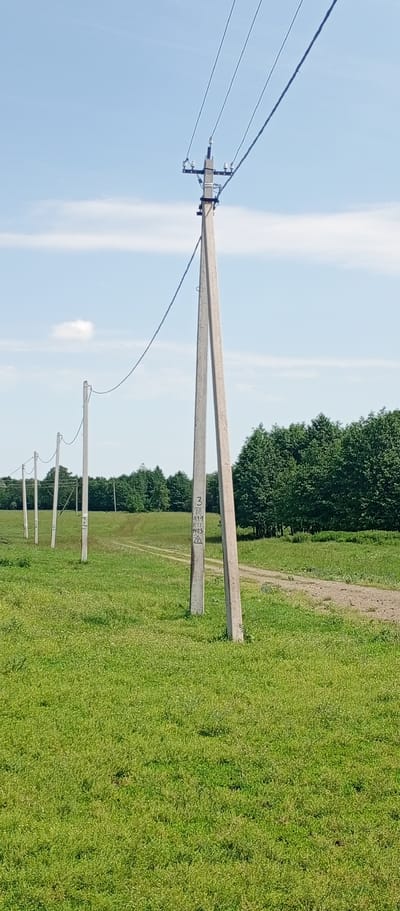 The image shows a rural landscape in Khusainovo, Russia, under a bright, partly cloudy sky. In the foreground, a row of concrete utility poles extends into the distance, with power lines strung between them. The poles are spaced evenly across a lush green meadow. A dirt road winds through the grass, leading towards a dense line of dark green trees that forms the horizon. One of the utility poles in the foreground has markings on it, including a triangle symbol and numbers. The overall scene depicts a serene, natural environment with signs of human infrastructure.