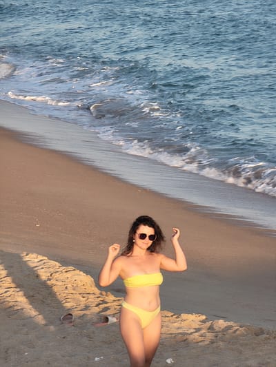 A young woman with dark, wavy hair and sunglasses stands on a sandy beach. She is wearing a yellow bikini and has her arms raised, with her hands near her head. The ocean is to her right, with gentle waves washing onto the shore. The sand has shadows cast across it, suggesting the sun is low in the sky. A pair of flip-flops are visible in the sand to her left. The scene appears to be Quarteira, Portugal, given the context provided.