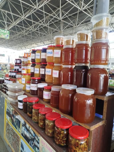This media file captures a vibrant marketplace scene, likely in Tashkent, Uzbekistan, showcasing a large display of honey jars and bee pollen. The setting is a covered market with a distinctive metal roof structure overhead. Sunlight filters through, suggesting it is daytime.
The primary subjects are an array of glass jars filled with various types of honey, distinguished by color and some by labels. There are also clear plastic containers filled with golden-yellow bee pollen stacked atop some of the honey jars. The jars are arranged on tiered wooden shelves and a long counter, creating a colorful and abundant display. Some jars contain whole nuts and dried fruits mixed with honey.
Visible text on the labels is predominantly in Cyrillic script. Some discernible words and phrases include "КУРАЙ" (Kuray), "ПРОПША" (Propsha), "МАЙСКИЙ" (May Sky - possibly a type of honey), and "ЧЕСТВЕННЫЙ МЕД" (Honest Honey) or "НАТУРАЛЬНЫЙ МЕД" (Natural Honey). Other labels mention "ВАШЕГО ЗДОРОВЬЯ" (For Your Health). There's also a sign partially visible with the word "ДОСТАВКА" (Delivery).
In the background, elements of the market are visible, including other stalls and potentially people. One image on a display board shows people working with bees, hinting at the production of the honey. The overall impression is one of a bustling, well-stocked market, offering a variety of natural products. The abundance and presentation of the goods suggest a focus on health and natural sustenance.